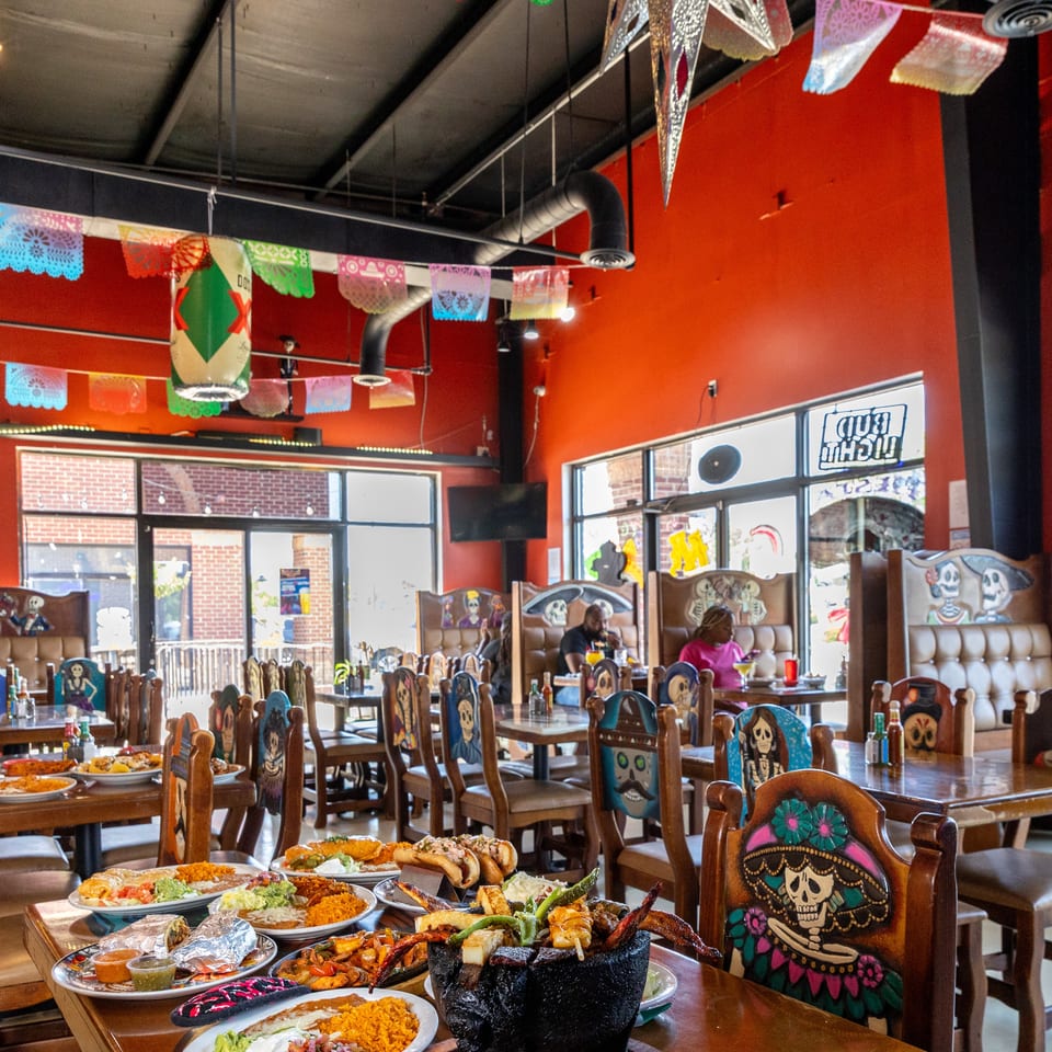 Colorful Mexican restaurant dining room with Day of the Dead chairs, papel picado decorations, diners, and plates of food.