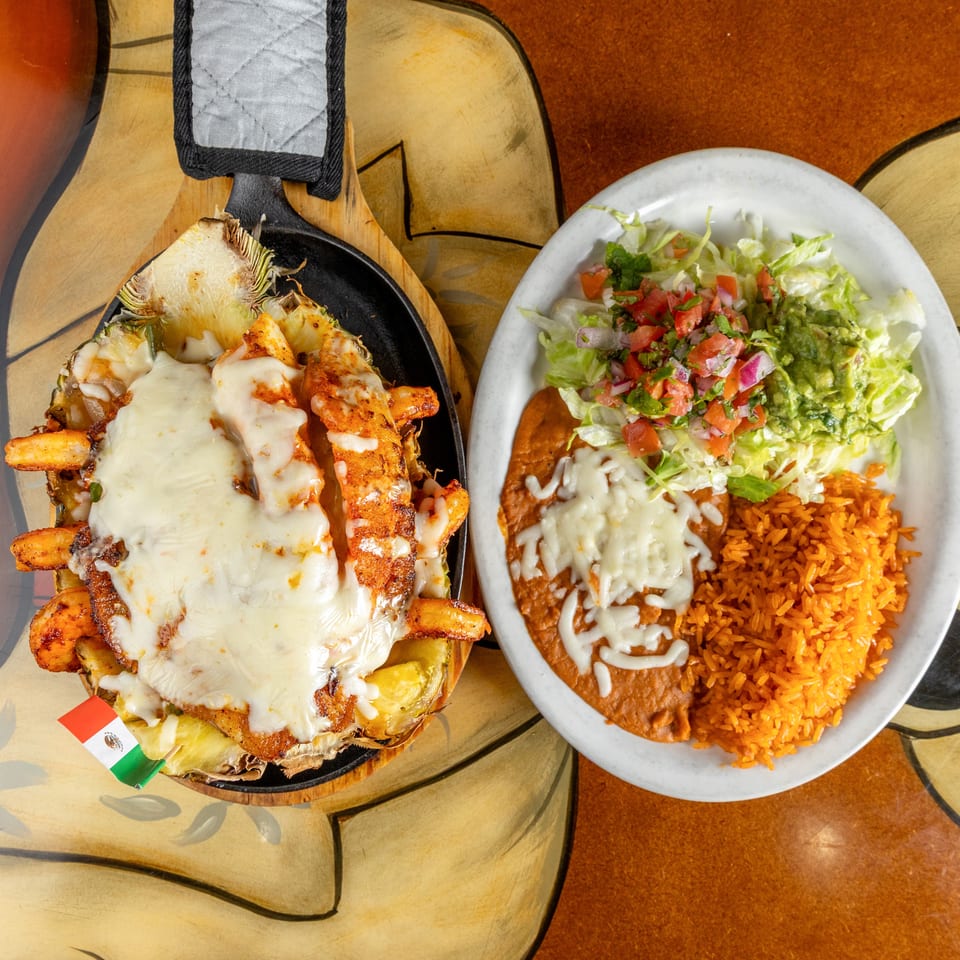 Mexican meal featuring cheesy grilled shrimp in a pineapple boat and a plate of rice, beans, lettuce, guacamole, and pico de gallo.