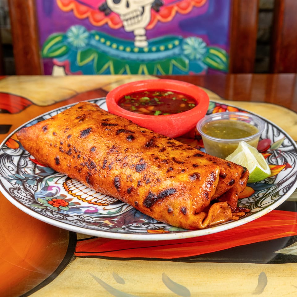 Two grilled, folded orange enchiladas on a colorful plate with red and green salsa, lime wedges, and a festive skull backdrop.