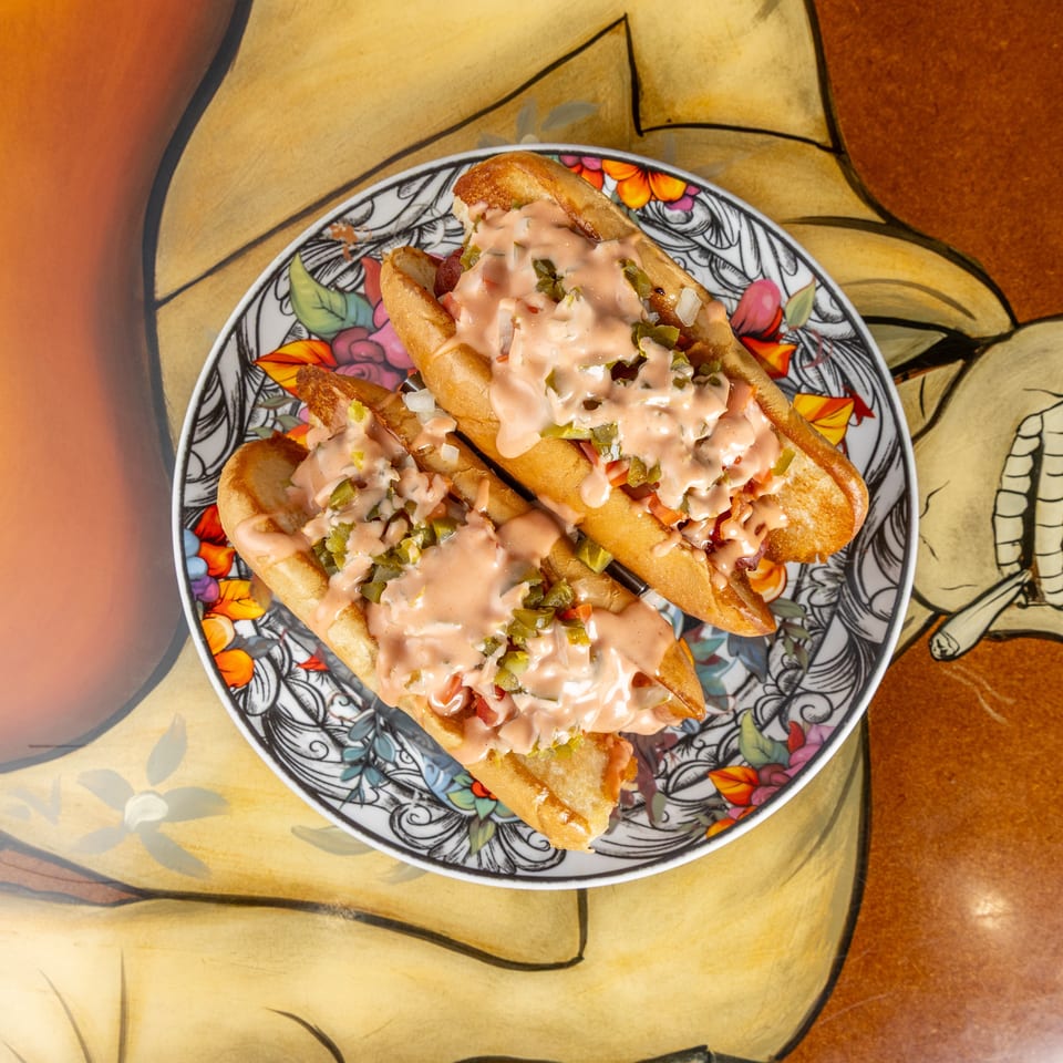 Loaded hot dogs topped with creamy pink sauce, chopped pickles, onions, and tomatoes on a colorful floral plate.