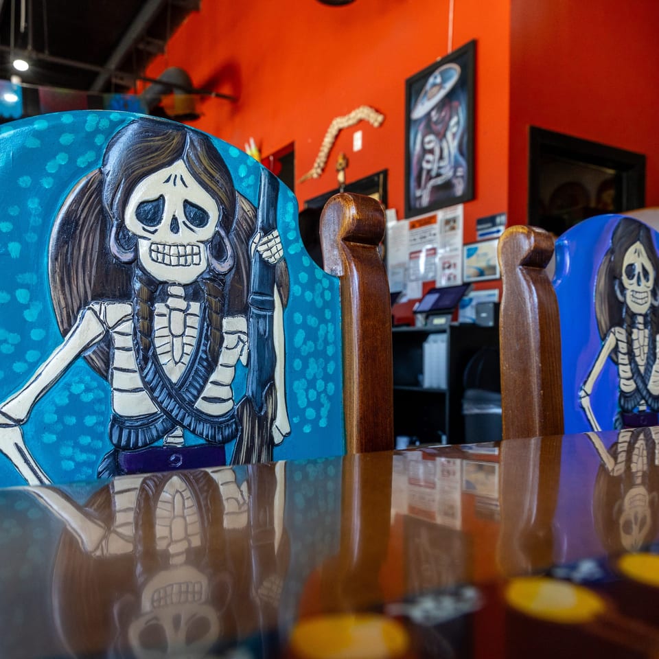 Colorful restaurant interior featuring painted skeletons on blue chairs, red walls, Mexican folk art, and reflective tables.