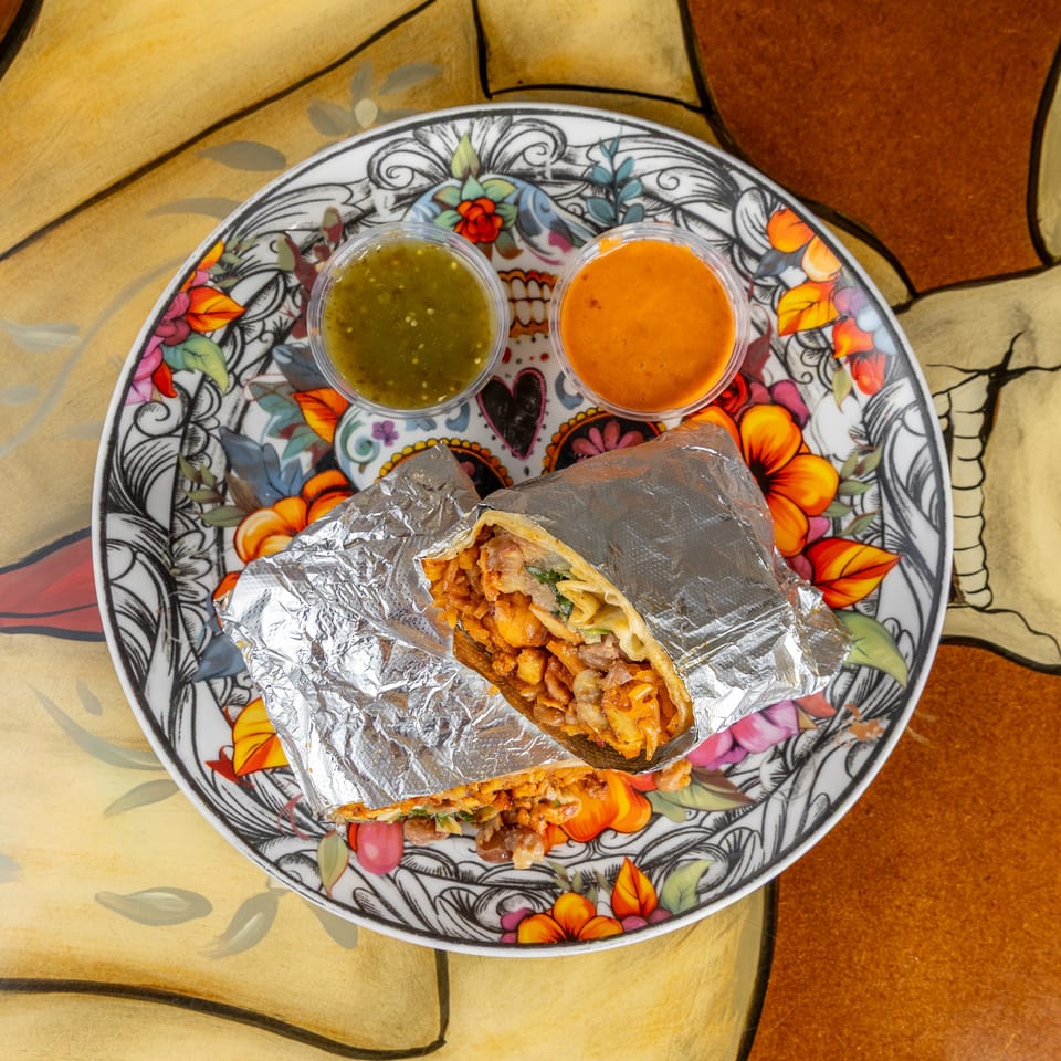 Foil-wrapped burrito cut open on a colorful skull-decorated plate, served with green and red salsa.