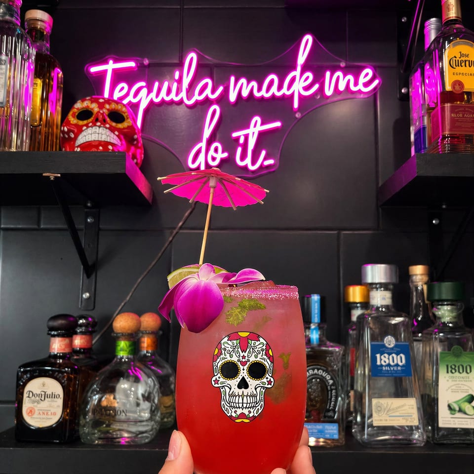 Hand holding a vibrant red cocktail with a sugar skull design, orchid, and mini umbrella, against a neon "Tequila made me do it" sign.