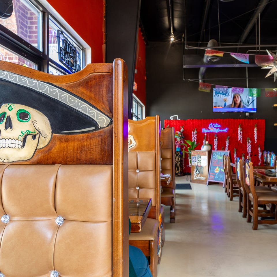 Colorful restaurant interior with skull-painted wooden booths, festive papel picado banners, television, and red floral wall decor.