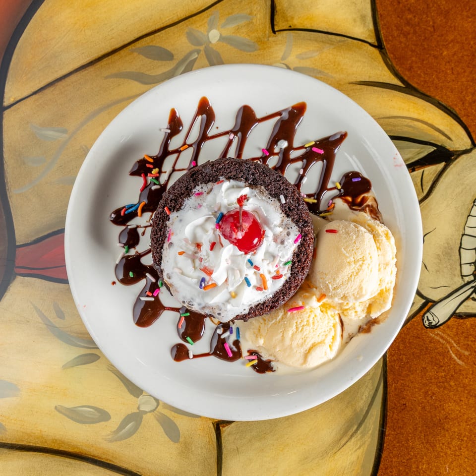Chocolate lava cake topped with whipped cream, sprinkles, and a cherry, served with vanilla ice cream and chocolate drizzle.