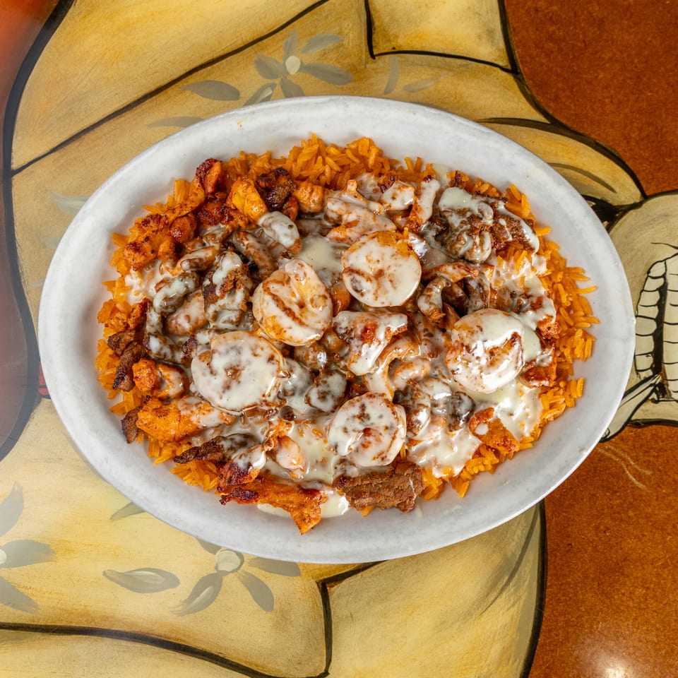 Plate of orange rice topped with grilled shrimp, chicken, steak, and creamy white sauce on a patterned table.