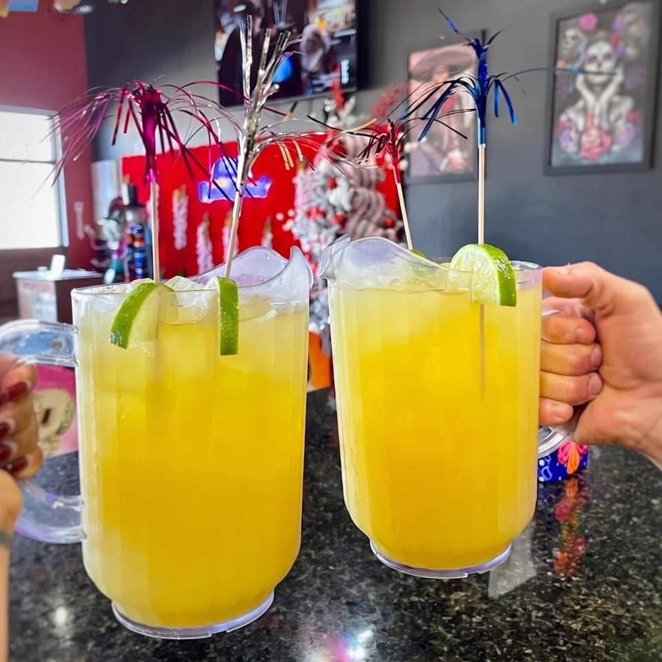 Two large pitchers of yellow margaritas with lime slices and festive stirrers on a dark counter.