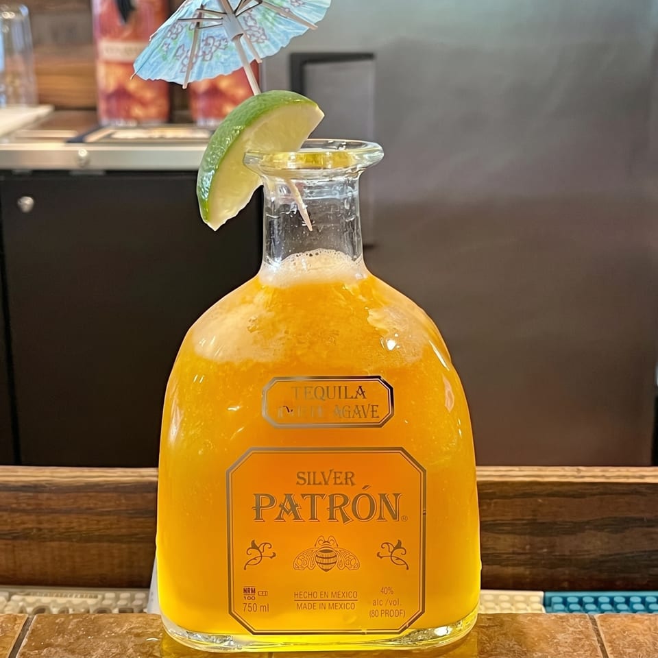 A Patron tequila bottle filled with a bright orange cocktail, garnished with a lime and paper umbrella.