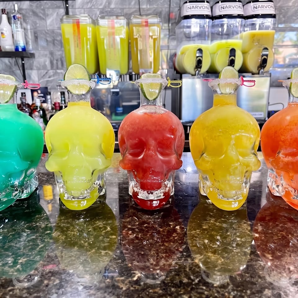 Five colorful skull-shaped margarita glasses with lime wedges and salted rims on a bar.