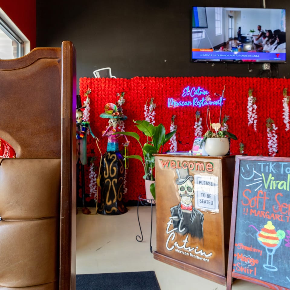 Colorful El Catrin Mexican restaurant entrance with red floral wall, neon sign, welcome board, chalkboard, and booth seating.