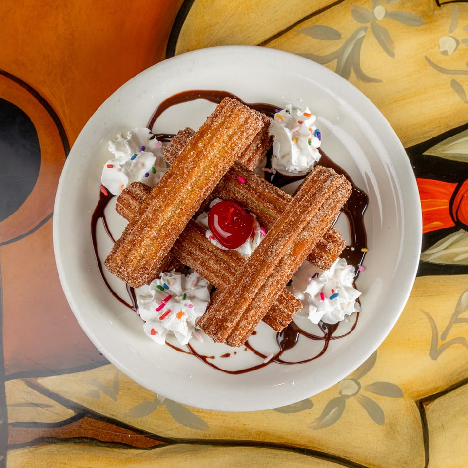 Cinnamon-sugar churros piled on a plate with whipped cream, rainbow sprinkles, cherry, and chocolate sauce.
