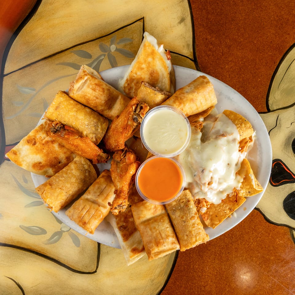 Platter of assorted fried appetizers with spring rolls, chicken wings, quesadillas, creamy toppings, and dipping sauces.