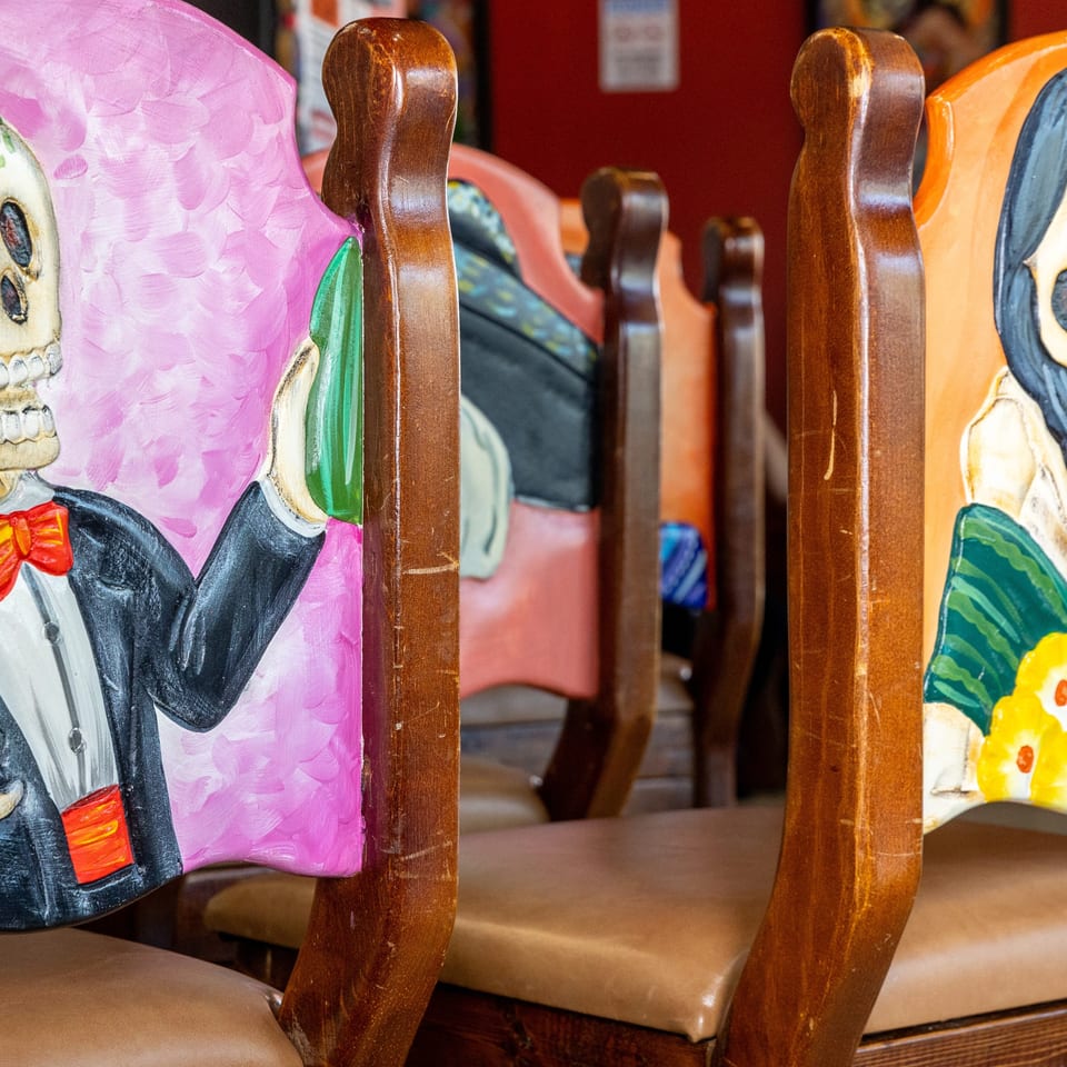 Colorful hand-painted skeleton chair backs with floral designs line a warmly lit restaurant dining area.