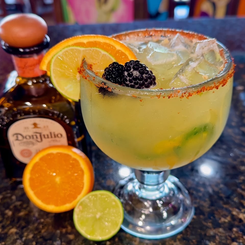 Citrus cocktail with blackberries, ice, and a chili-salt rim beside orange and lime slices on a restaurant bar.