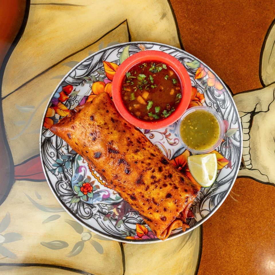 Crispy golden stuffed pastry served on a floral plate with red salsa, green sauce, cilantro, and lime.