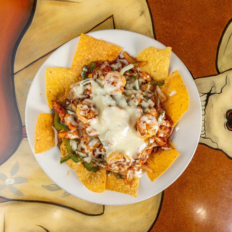 Loaded nachos topped with grilled shrimp, melted cheese, peppers, and onions on a white plate over a colorful table.