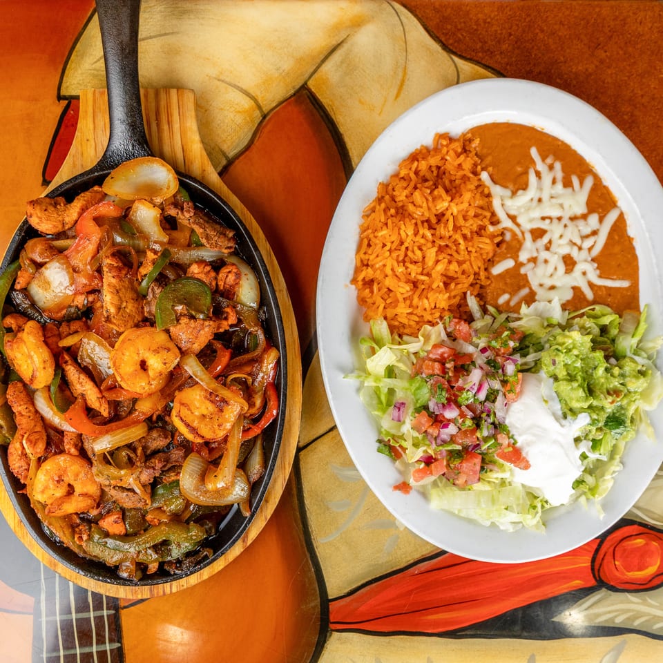 Mexican fajitas with shrimp, chicken, peppers, and onions served alongside rice, beans, salad, guacamole, and sour cream.
